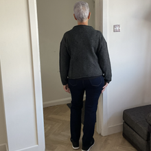 Load image into Gallery viewer, A person viewed from behind wearing a dark gray knit sweater and dark navy jeans, standing in a doorway inside a home with light-colored walls and wood flooring.