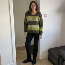 Load image into Gallery viewer, A woman stands in a doorway wearing a chunky knit sweater with horizontal stripes in olive green, yellow-green, and gray, paired with dark jeans and black boots.