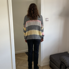 Load image into Gallery viewer, A person viewed from behind wearing a chunky knit striped sweater with gray, pink, yellow, and white bands, paired with dark jeans in an indoor room setting.