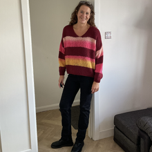Load image into Gallery viewer, A woman stands in a doorway wearing a knit sweater with horizontal stripes in burgundy, pink, orange, and yellow, paired with dark pants and black shoes.