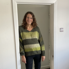 Load image into Gallery viewer, A woman with shoulder-length brown hair stands in a doorway wearing a chunky knit sweater with horizontal stripes in olive green, dark gray, and light yellow-green, paired with dark pants.