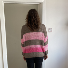 Load image into Gallery viewer, A person viewed from behind wearing a chunky knit sweater with horizontal stripes in taupe, light pink, and magenta, paired with dark jeans, standing in a doorway.