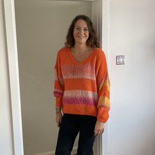 Load image into Gallery viewer, A woman wearing a bright orange knit sweater with pink and yellow horizontal stripes, paired with dark jeans, standing in a doorway against a white wall.