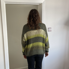 Load image into Gallery viewer, A person viewed from behind wearing a chunky knit sweater with horizontal stripes in olive green, beige, and yellow-green, standing in a doorway.
