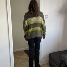 Load image into Gallery viewer, A person viewed from behind wearing a loose, fuzzy striped sweater with olive green, beige, and yellow-green bands, paired with dark jeans and black boots, standing in a doorway inside a home.