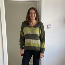 Load image into Gallery viewer, A woman wearing a chunky knit striped sweater with olive green, lime green, and cream horizontal bands, standing in a doorway.