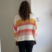 Load image into Gallery viewer, A person viewed from behind wearing a chunky knit sweater with horizontal stripes in cream, yellow, orange, and pink, paired with dark jeans, standing in a doorway.