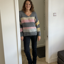 Load image into Gallery viewer, A woman wearing a chunky knit V-neck sweater with horizontal stripes in gray, pink, cream, and yellow, paired with black pants and boots, standing in a doorway of a home interior.