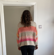 Load image into Gallery viewer, A person viewed from behind wearing a chunky knit sweater with horizontal stripes in beige, orange, pink, and yellow, standing in a doorway.