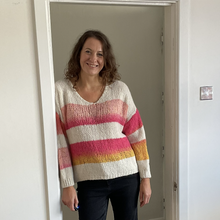Load image into Gallery viewer, A woman smiling and standing in a doorway wearing a chunky knit sweater with horizontal stripes in cream, pink, peach, and yellow, paired with dark jeans.
