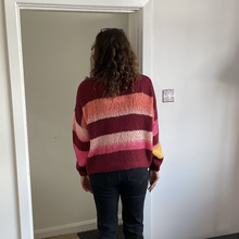 Load image into Gallery viewer, A person standing with their back to the camera wearing a maroon and pink striped knit sweater with peach and yellow stripes, paired with dark jeans, in an indoor setting.
