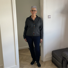 Load image into Gallery viewer, An older woman with short gray hair and glasses stands in a doorway wearing a dark gray knit cardigan with colorful buttons, dark navy pants, and black sneakers in a home setting.