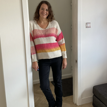 Load image into Gallery viewer, A woman stands in a doorway wearing a chunky knit sweater with horizontal stripes in cream, pink, orange, and yellow, paired with dark jeans and black shoes.