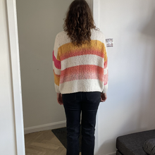 Load image into Gallery viewer, A person viewed from behind wearing a fuzzy knit striped sweater with horizontal bands of cream, yellow, orange, and pink, paired with dark jeans in an indoor setting.