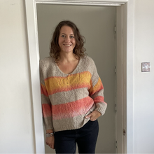 Load image into Gallery viewer, A woman smiling and standing in a doorway wearing a chunky knit sweater with horizontal stripes in beige, yellow, orange, and pink, paired with dark blue jeans.