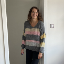 Load image into Gallery viewer, A woman wearing a chunky knit V-neck sweater with horizontal stripes in gray, pink, beige, and yellow, standing in a doorway.