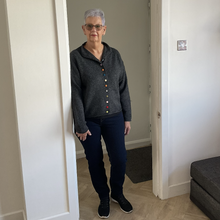 Load image into Gallery viewer, An older woman with short gray hair and glasses stands in a doorway wearing a dark gray knit zip-up cardigan with colorful buttons, dark blue jeans, and black sneakers.