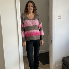 Load image into Gallery viewer, A woman wearing a chunky knit sweater with horizontal stripes in brown, light pink, and magenta, paired with dark jeans and black boots, standing in a doorway inside a home.