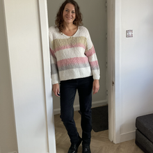 Load image into Gallery viewer, A woman stands in a doorway wearing a chunky knit sweater with horizontal stripes in cream, beige, pink, and gray, paired with dark jeans and black boots.

