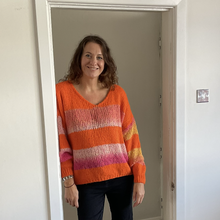 Load image into Gallery viewer, A woman with curly brown hair stands in a doorway wearing a bright orange knit sweater with horizontal stripes in pink, white, and yellow, paired with dark pants.
