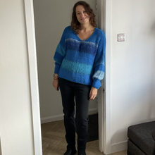 Load image into Gallery viewer, A woman wearing a blue and teal striped fuzzy knit sweater, black pants, and black boots, standing in a doorway of a home interior.
