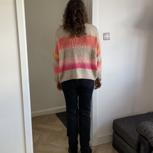 Load image into Gallery viewer, A person viewed from behind wearing a chunky knit sweater with horizontal stripes in beige, orange, pink, and purple, paired with dark jeans, standing in a doorway inside a home.
