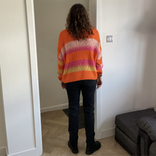 Load image into Gallery viewer, A person viewed from behind wearing a vibrant orange knit sweater with pink, purple, and yellow horizontal stripes, paired with dark jeans and black boots, standing in a doorway.
