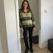 Load image into Gallery viewer, A woman stands in a doorway wearing a chunky knit sweater with horizontal stripes in olive green, dark gray, and light yellow-green, paired with dark jeans and black shoes.

