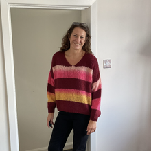 Load image into Gallery viewer, A woman wearing a maroon, pink, and yellow striped knit sweater and dark jeans, standing in a doorway of a white-walled room.
