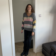 Load image into Gallery viewer, A woman stands in a doorway wearing a chunky knit V-neck sweater with horizontal stripes in gray, pink, yellow, and white, paired with black pants and black shoes.
