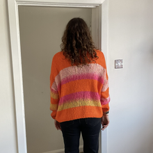 Load image into Gallery viewer, A person viewed from behind wearing a chunky knit sweater with horizontal stripes in orange, pink, purple, and yellow, paired with dark jeans, standing in a doorway.
