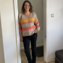 Load image into Gallery viewer, A woman stands in a doorway wearing a fuzzy knit sweater with horizontal stripes in beige, orange, yellow, and pink, paired with dark pants and black boots.
