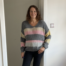 Load image into Gallery viewer, A woman wearing a chunky knit sweater with horizontal stripes in gray, cream, pink, and yellow, paired with dark jeans, standing in a doorway.
