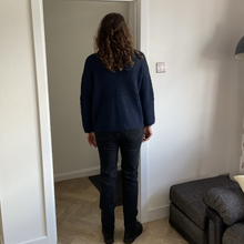 Load image into Gallery viewer, A person with long curly brown hair stands with their back to the camera, wearing a dark navy blue knit sweater and dark jeans, positioned in front of an open white doorway in a home interior.
