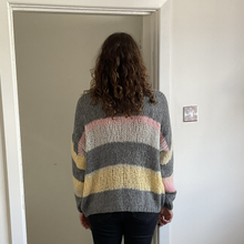 Load image into Gallery viewer, A person viewed from behind wearing a chunky knit striped sweater with horizontal bands of gray, white, pink, and pale yellow, standing in a doorway.
