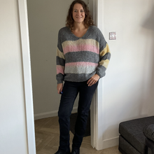 Load image into Gallery viewer, A woman wearing a chunky knit sweater with horizontal stripes in gray, yellow, and pink, paired with dark jeans and black boots, standing in a doorway of a home interior.
